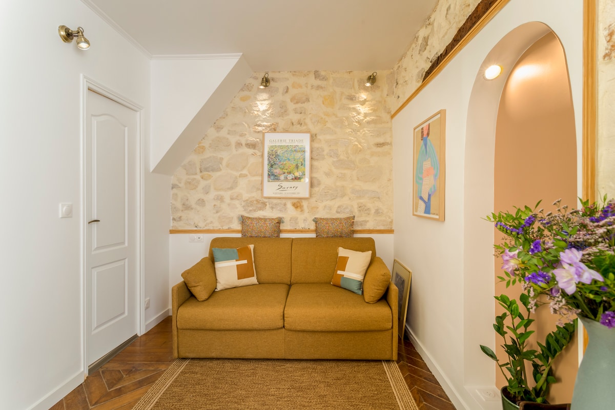 A cozy living area features a comfortable brown sofa adorned with colorful cushions. The stone walls add texture, while warm lighting highlights the space. A woven area rug sits beneath, and a small decorative archway leads to another room. Fresh flowers are displayed in a vase.