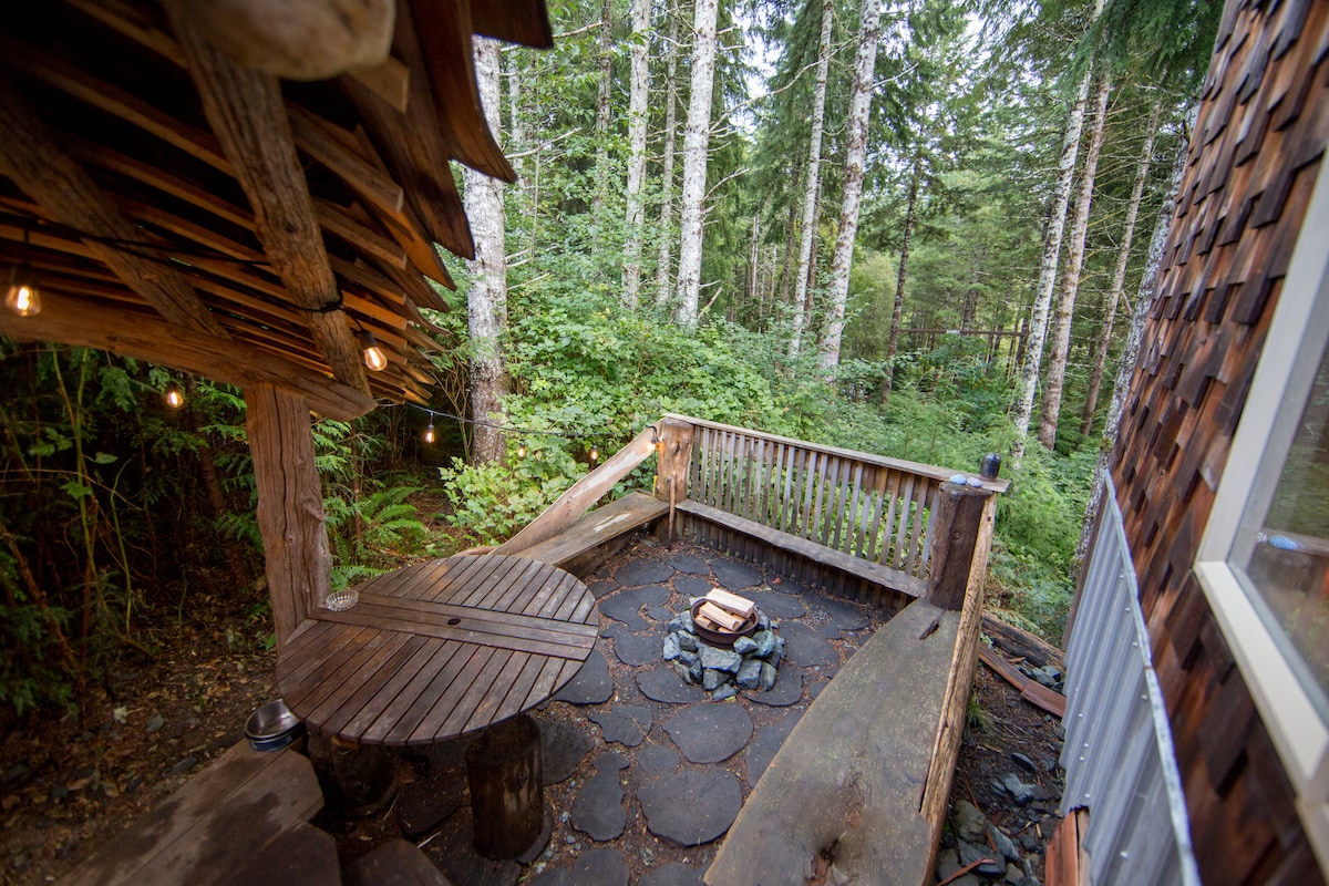 An outdoor seating area features a round wooden table surrounded by natural stone. A fire pit is positioned at the center, with wooden benches complementing the rustic setting. Lush greenery and towering trees provide a secluded backdrop, enhancing the peaceful atmosphere.