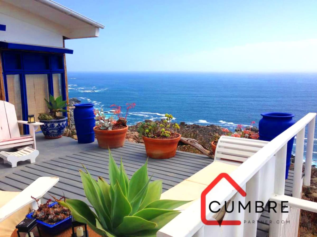 A sunlit deck overlooks the ocean, featuring potted plants and vibrant blue containers. Two white chairs provide seating to enjoy the coastal view, while the surrounding landscape showcases a mix of natural textures and colors under a clear blue sky.