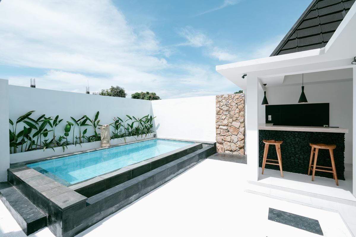 A private pool is surrounded by smooth stone and white walls, complemented by tropical plants along one side. An outdoor bar area features two wooden stools and a minimalist counter, with a clear blue sky above and bright sun reflecting off the water.