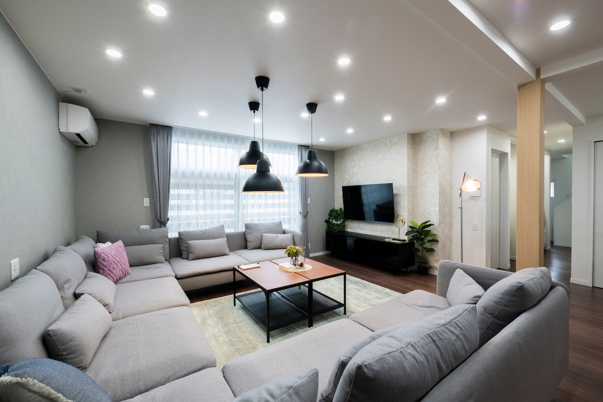 A spacious living room features soft gray sectional sofas arranged around a central wooden coffee table. Soft lighting is provided by pendant lamps above. Large windows with sheer curtains allow natural light to fill the area, complemented by decorative plants and a television mounted on the wall.