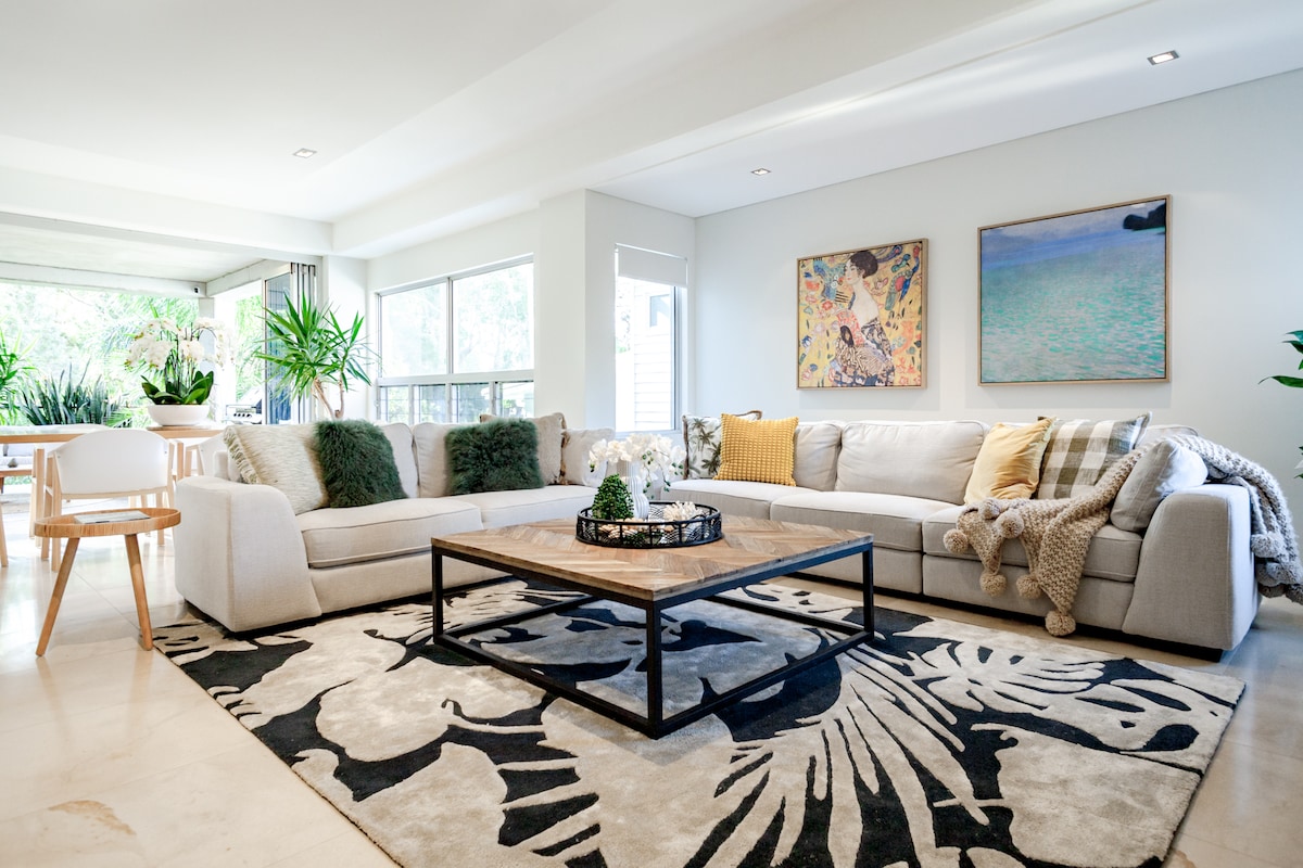 The living area features a spacious, light-filled layout, furnished with a large sectional sofa and a coffee table atop a patterned rug. Framed artwork adorns the walls, complemented by greenery from indoor plants, creating a relaxing and inviting space.
