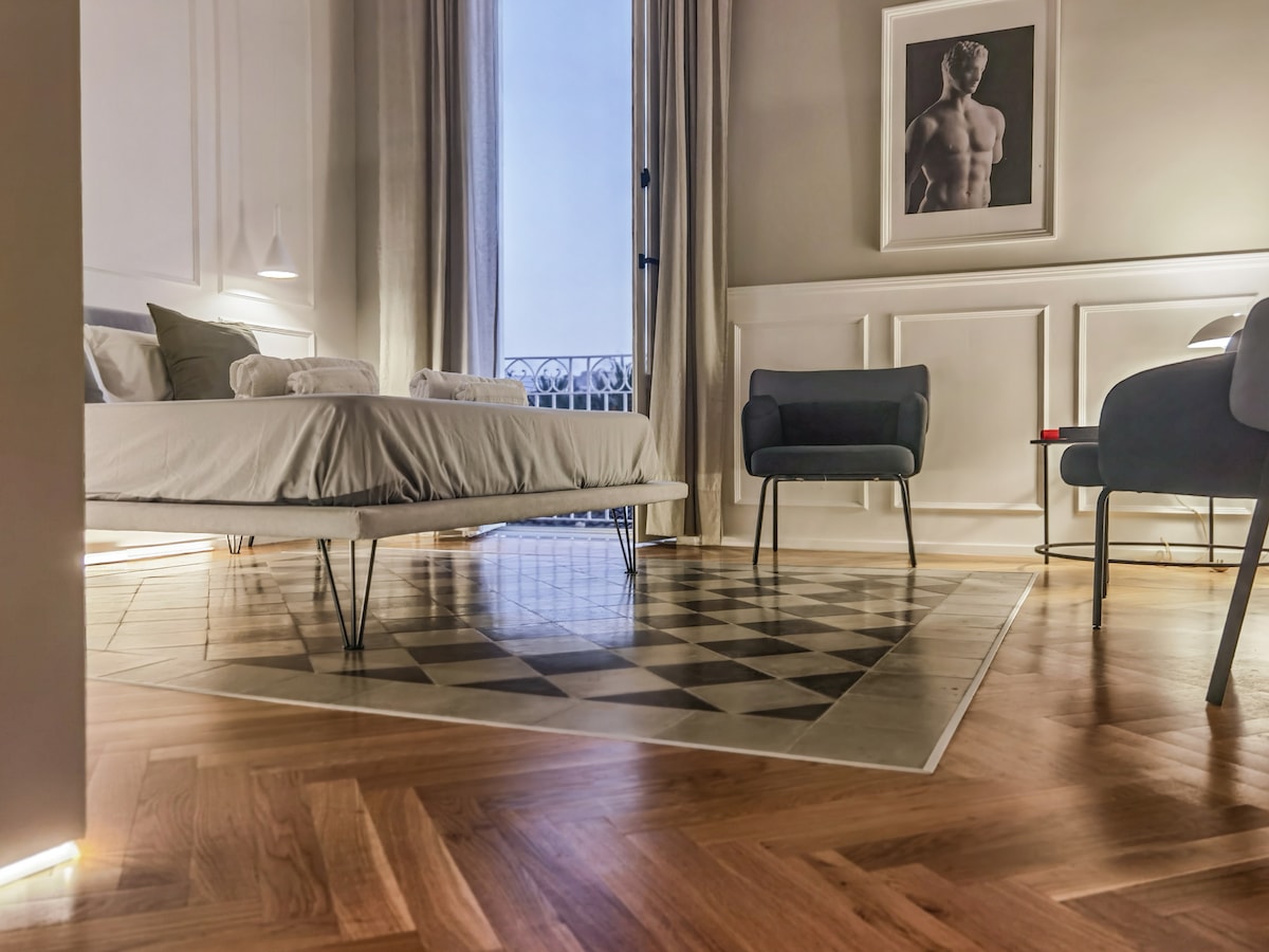 A serene bedroom is presented with a spacious bed, flanked by stylish blue armchairs. A sleek wooden floor with a geometric rug adds warmth. Natural light streams in through large windows, enhancing the calm ambiance. A framed artwork complements the elegant decor.