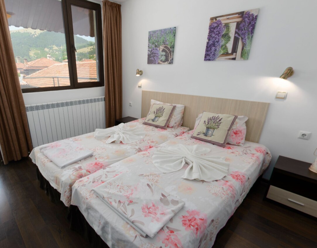 A well-appointed sleeping area is furnished with two spacious beds, each adorned with floral-patterned bedding. Accents of lavender artwork are displayed on the walls. Natural light enters through a large window, offering views of the surrounding mountains and town.