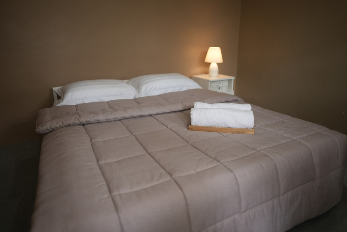 The sleeping area features a spacious bed covered with a soft, light-colored quilt. A neatly arranged white towel rests on a wooden tray beside the bed, while a bedside lamp casts a subtle glow against the warm brown walls.