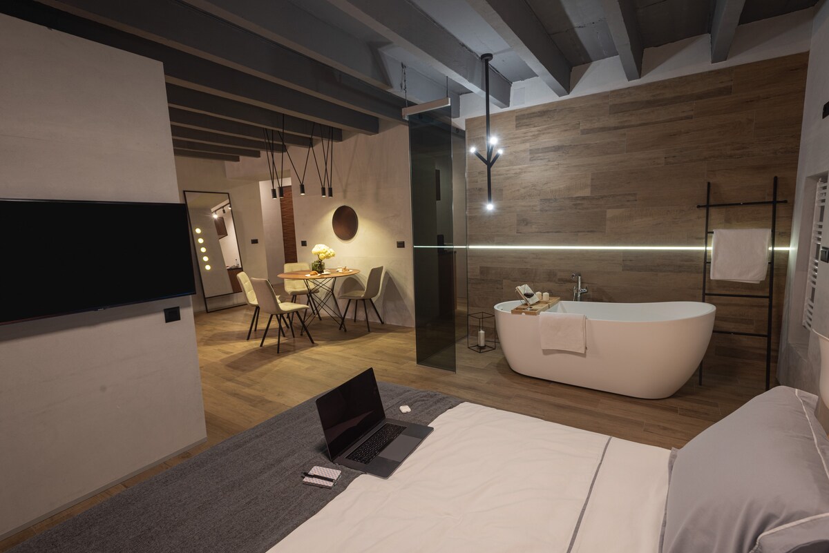 An open-plan living space is shown, featuring a spacious bed with a gray throw. A freestanding bathtub is positioned against a wooden wall, while a dining area with four chairs is visible in the background. A sleek television is mounted on the adjacent wall.