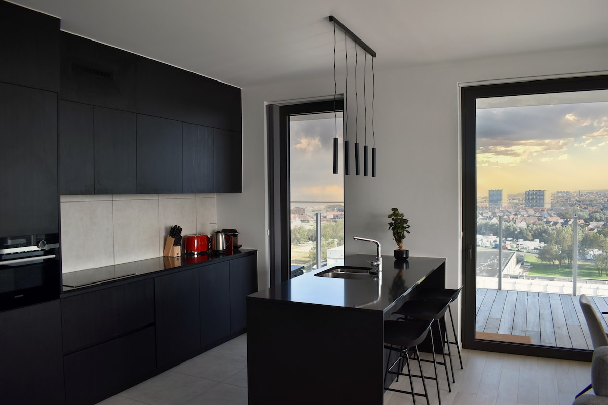 A modern kitchen is equipped with sleek black cabinetry and a central island with seating for four. Large windows offer a panoramic view of the city, while natural light brightens the space. Essential appliances and cooking utensils are neatly arranged.