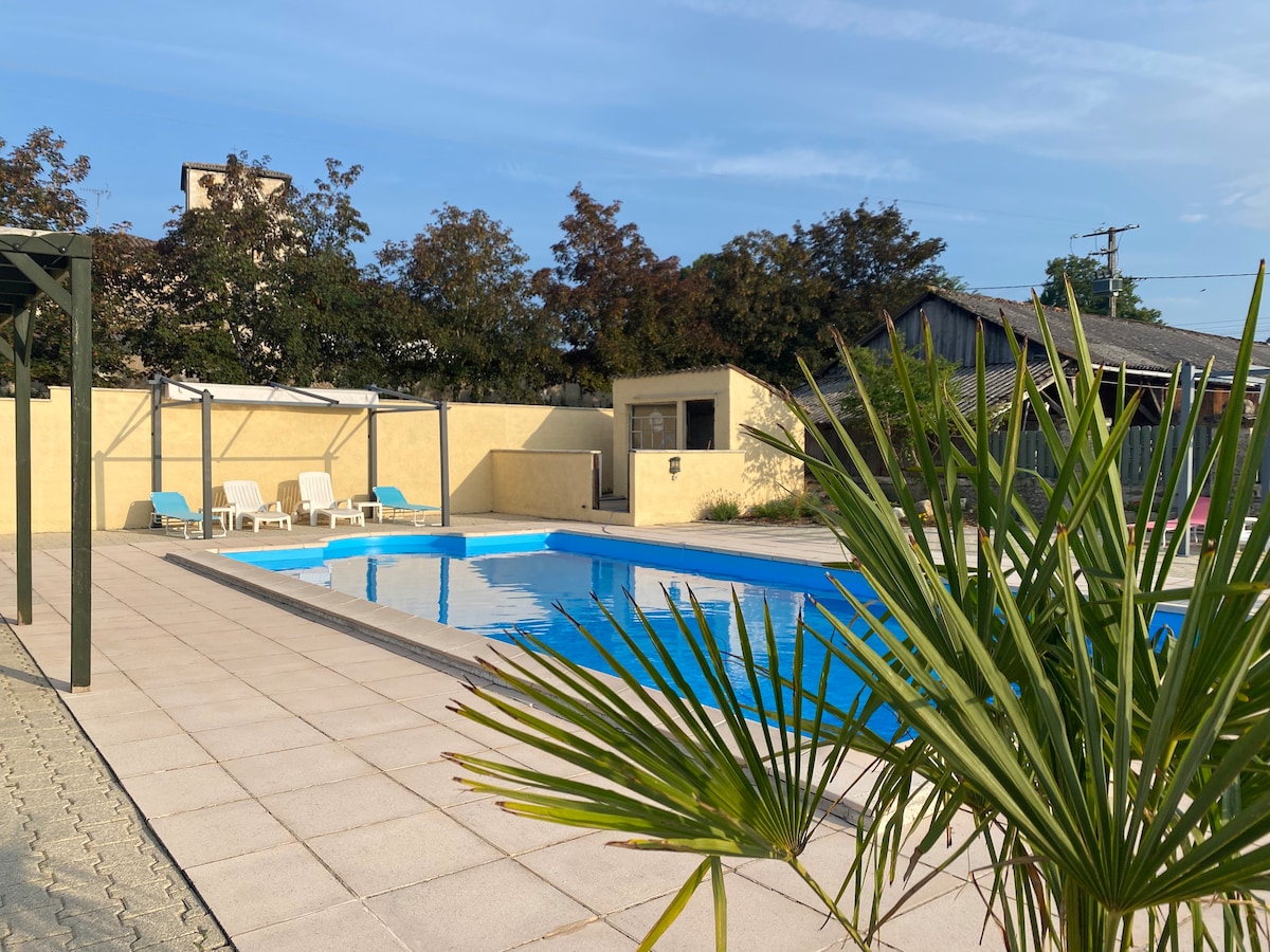 A large saltwater pool is framed by a spacious patio area. Several sun loungers and chairs are positioned nearby, inviting relaxation. Lush green plants add a natural touch to the serene setting, while a fence and building are visible in the background.