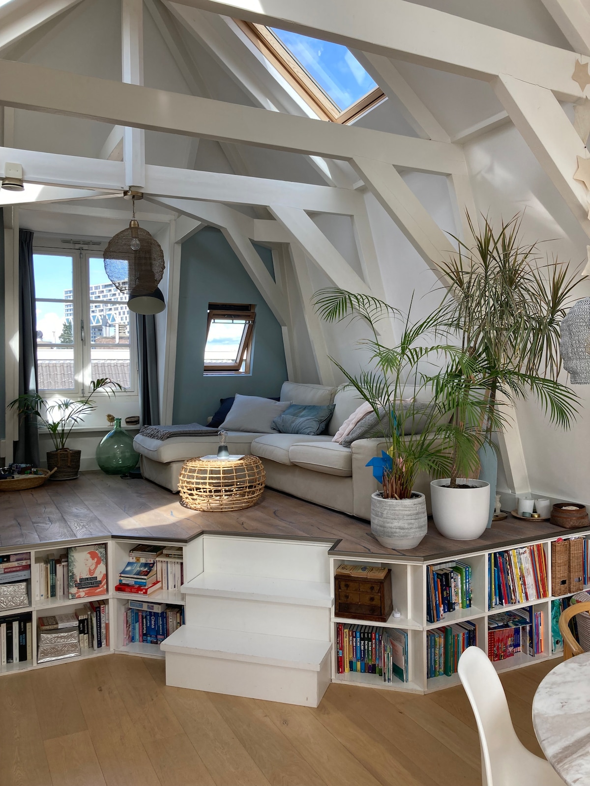 A bright living area showcases a spacious sofa, surrounded by potted plants. Natural light is captured through skylights, illuminating the wooden floor. Steps lead to a low bookshelf filled with various books, creating an inviting space with muted blue and white accents.
