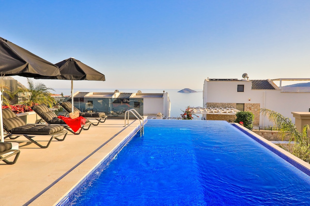 An infinity pool glistens under the sun, featuring vibrant blue water. Sun loungers with umbrellas are positioned nearby, offering a relaxing space. The serene view extends over Kalamar Bay, with distant islands enhancing the picturesque backdrop.