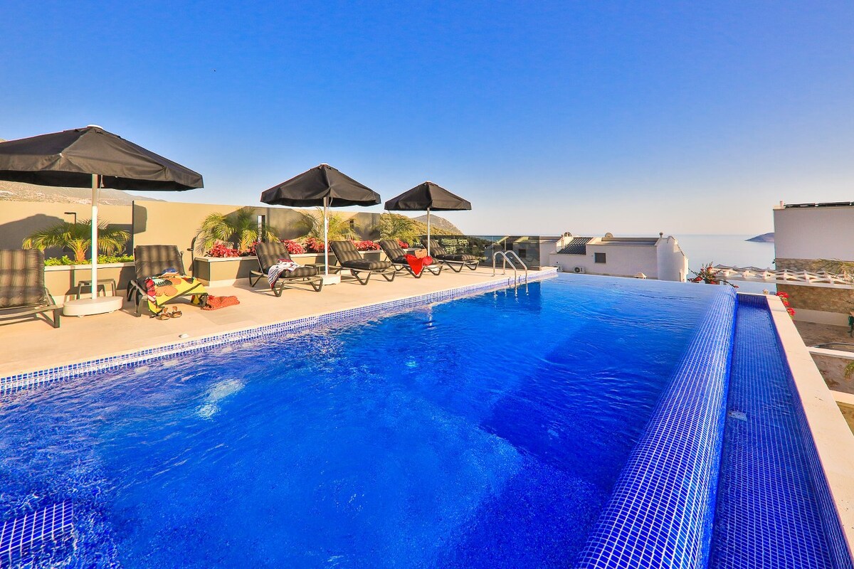 An infinity pool captures the expansive views of Kalamar Bay and the islands. Four black sun umbrellas provide shade over lounge chairs placed along the poolside, contributing to a serene outdoor space under a clear blue sky.