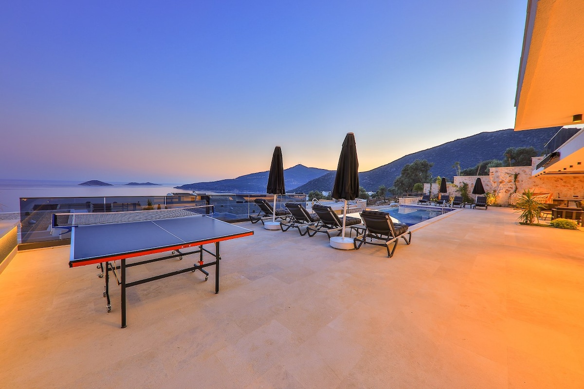 An outdoor terrace offers stunning views of the surrounding hills and Kalamar Bay. Sun loungers with black umbrellas are arranged along the edge, while a ping pong table is positioned centrally on the spacious patio, providing a recreational area for guests.