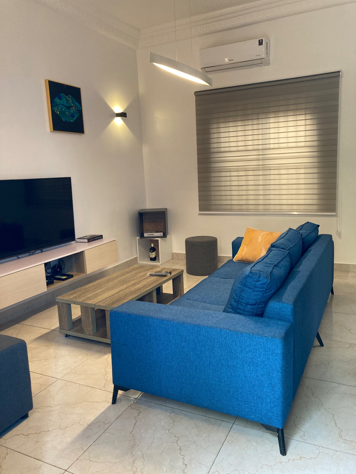 A modern living area is presented, featuring a blue sofa with cushions, a flat-screen TV mounted on a light-colored media console, and a wooden coffee table. Soft lighting highlights the room, while a window with shades allows natural light to enter.