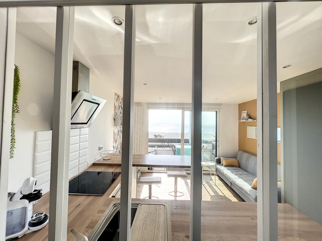 The open-plan living area features a view of the sea through large glass doors. A modern kitchen is equipped with essential appliances, while a dining table and cozy seating area complete the space. Soft light streams in, enhancing the inviting atmosphere.