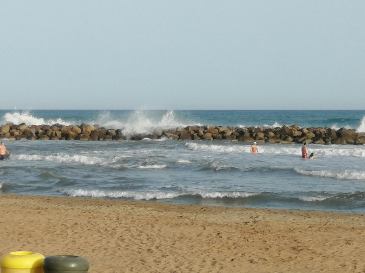 The image captures a serene beach scene, featuring gentle waves lapping against a rocky jetty. A few individuals can be seen enjoying the water, with soft sand stretching along the shoreline under a clear sky.