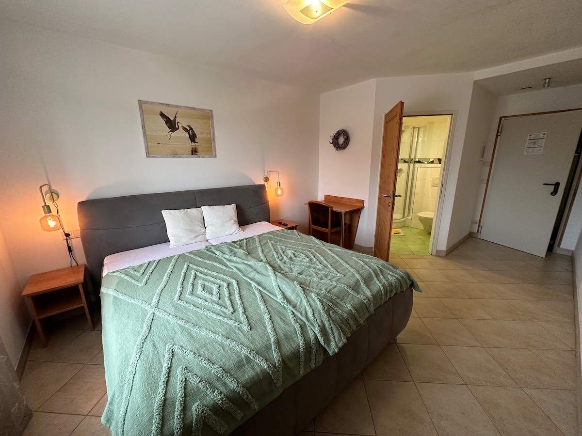 A spacious bedroom features a large bed with a green blanket, complemented by two bedside tables. Soft lighting is provided by wall-mounted lamps. A piece of wall art depicting birds adds a serene touch, while a bathroom door is visible in the background.