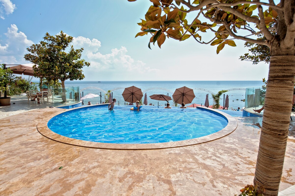 A circular pool is surrounded by a smooth, tiled deck with lounge chairs and umbrellas scattered around. Lush green trees add natural elements to the space, while the tranquil sea and sky complete the scenic view in the background.