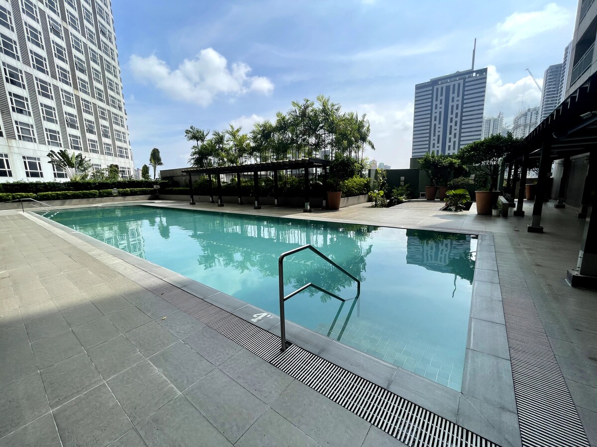 A clear and spacious swimming pool is visible, surrounded by greenery and potted plants. Sunlight reflects off the water's surface, and modern high-rise buildings form the backdrop.