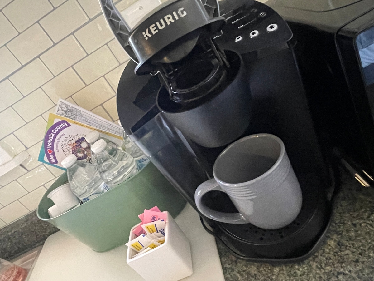 A compact kitchenette features a Keurig coffee maker alongside a gray mug positioned on the drip tray. A green container holds bottled water, while a small caddy with sweeteners is present. The countertops are lined with white subway tiles, enhancing the functional space.