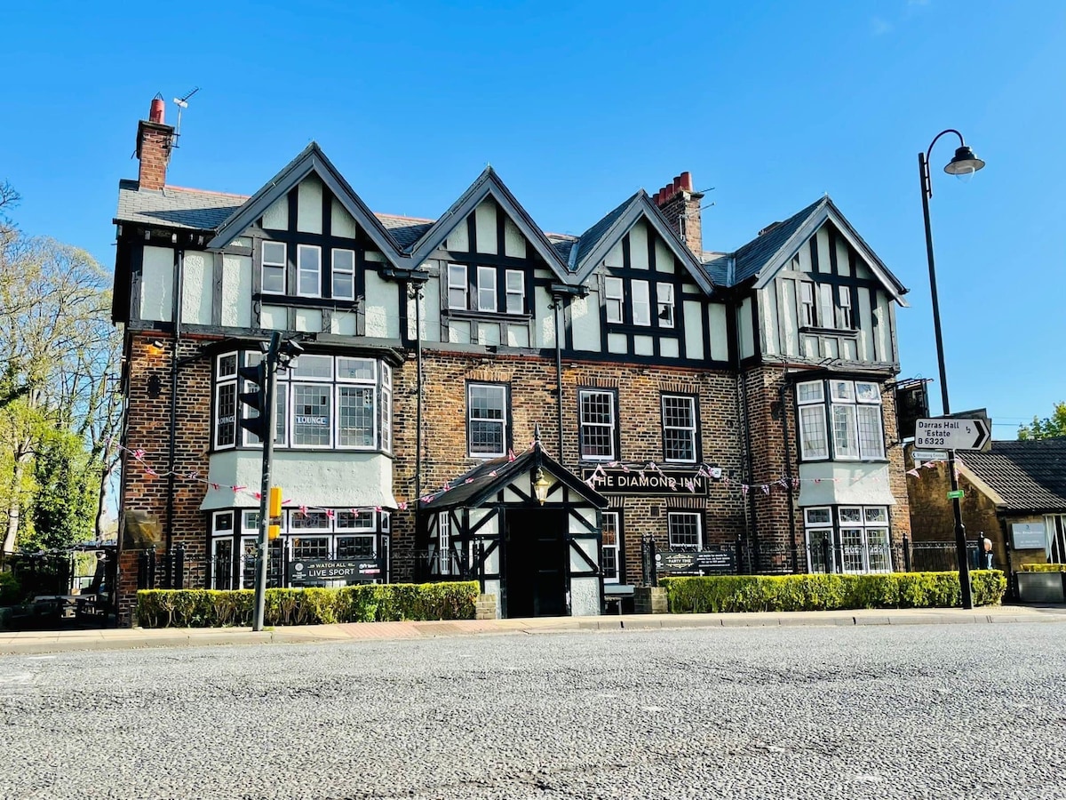 The three-story inn features a traditional design with timber framing and stone accents. Large windows create a bright facade, while a distinctive entrance is framed by greenery. The building is positioned near the road, with a clear view of the welcoming public house signage.