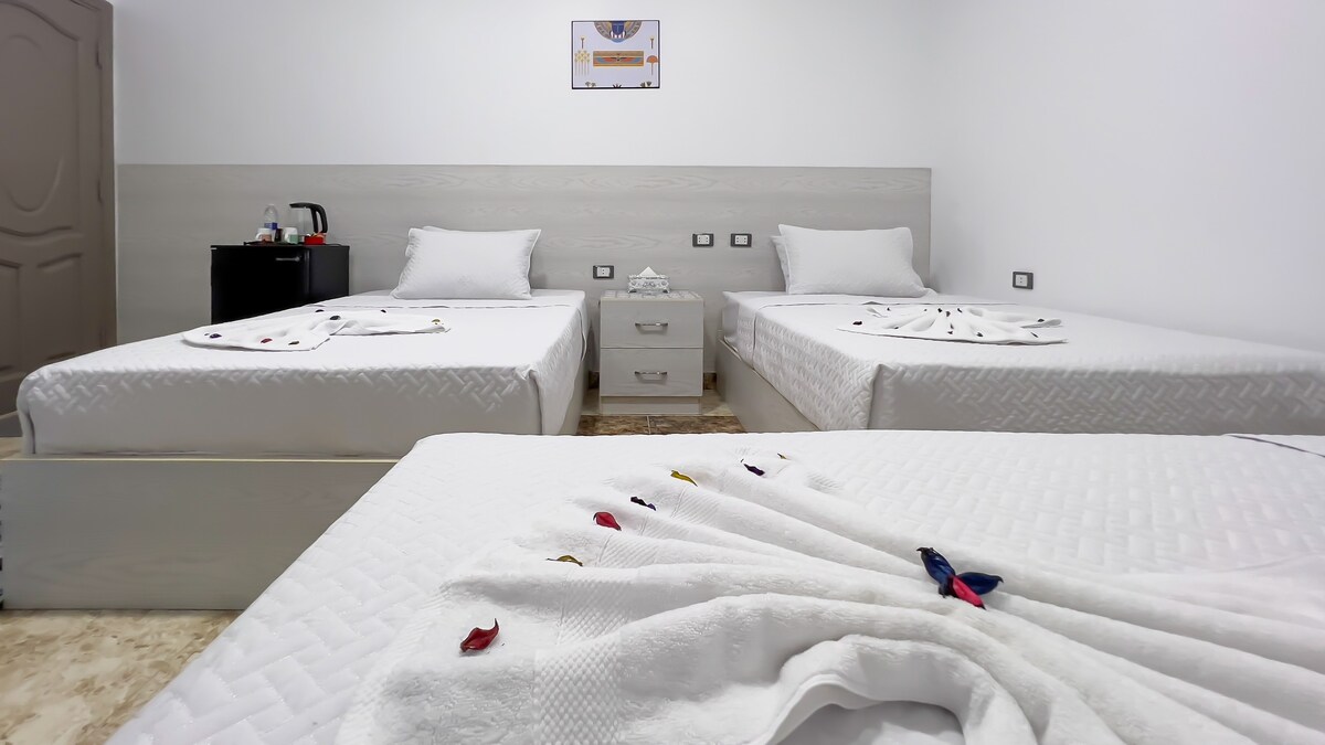 A clean and well-lit guest room features multiple beds with crisp white linens and decorative towels arranged neatly. A small kitchenette area with a kettle is visible, alongside bedside tables. A minimalistic design and light-colored walls contribute to a fresh ambiance.