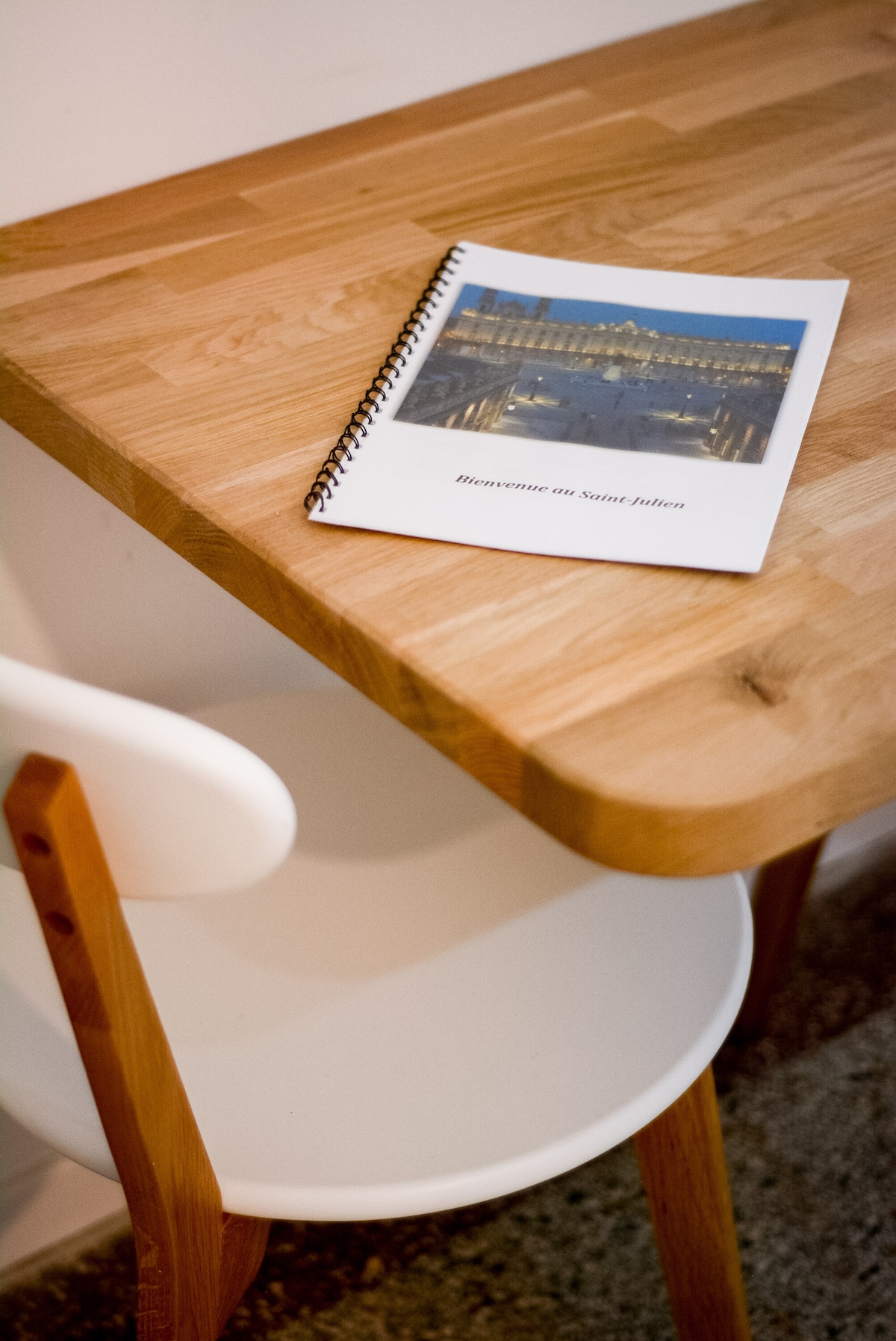 A wooden table is visible, featuring a rounded edge and a simple design. A spiral-bound booklet rests on the surface, displaying an image of a well-lit waterfront scene. A white chair with wooden legs complements the table.