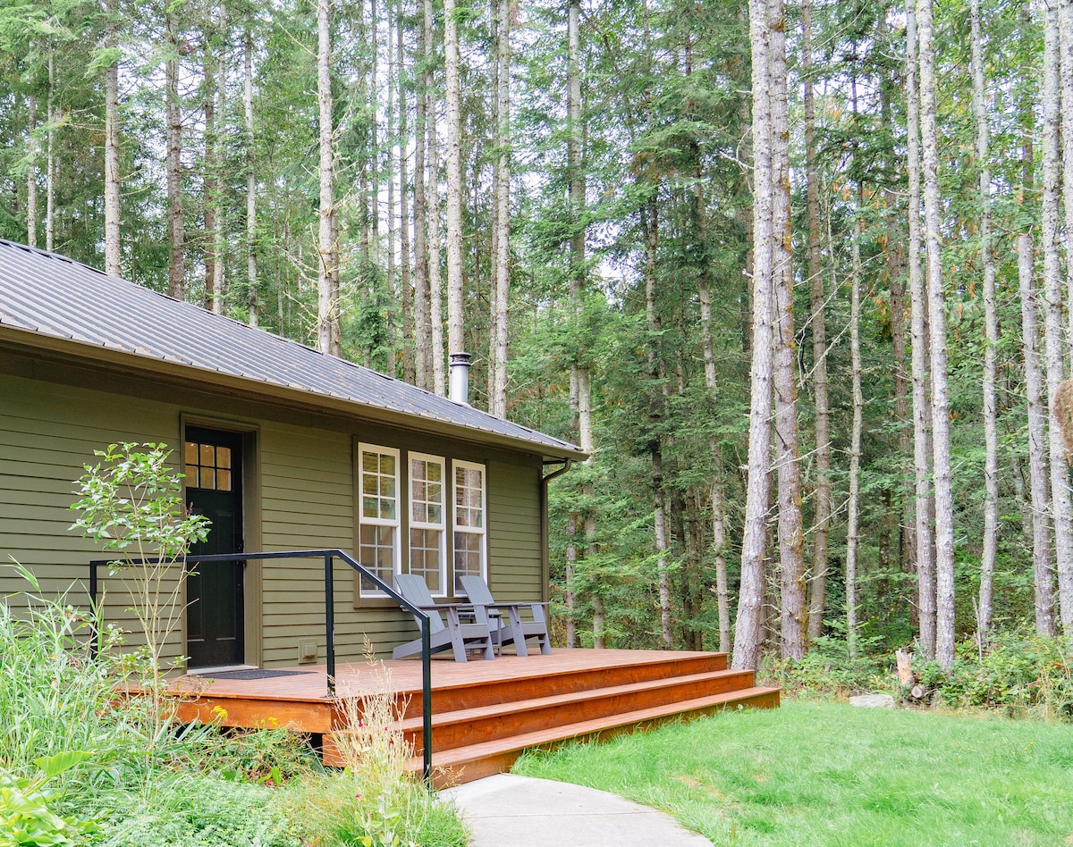 A charming guesthouse nestled among tall trees. The exterior showcases a wooden deck with two chairs positioned for relaxation. Large windows allow natural light to illuminate the indoors, while a stone path leads to the entrance, surrounded by lush greenery.