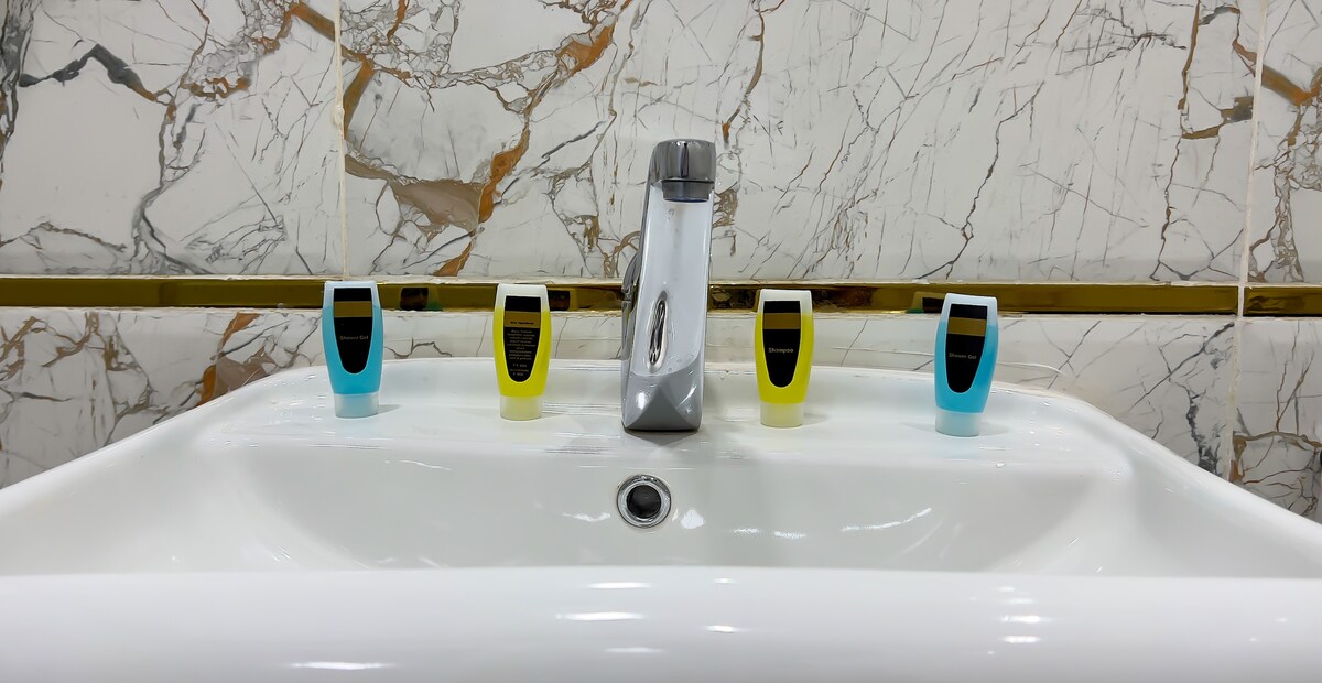A modern bathroom sink features a sleek chrome faucet. Multiple dispensers for soap or lotion are neatly arranged on the sink, each marked with clear labels. The backdrop is adorned with stylish marble, creating a clean and sophisticated atmosphere.