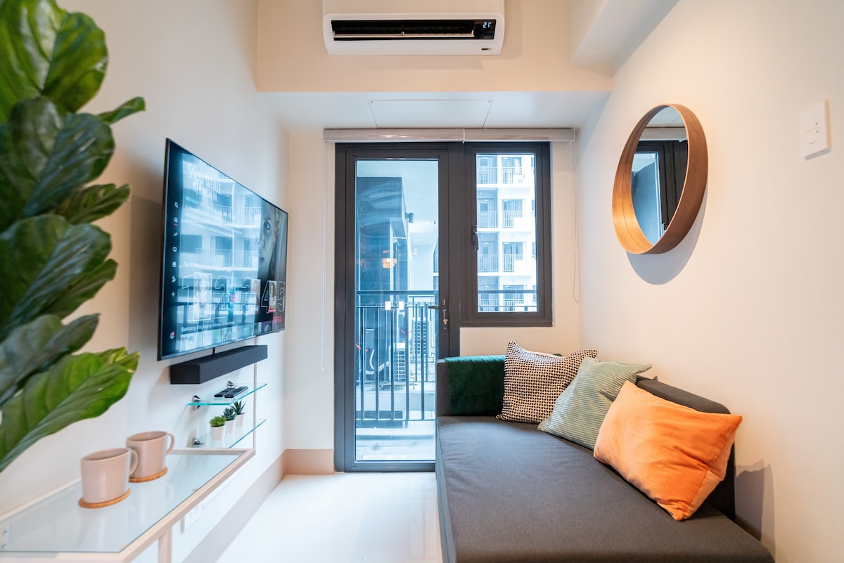 A compact living area features a gray sectional sofa adorned with decorative pillows in various colors. A flat-screen television is mounted on the wall opposite, with a small side table holding two cups. Natural light filters through the sliding glass doors leading to a balcony.