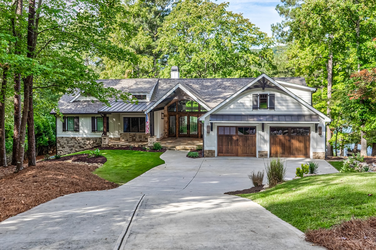 The exterior of a newly built lakefront home is visible, featuring a blend of stone and wood accents. A spacious driveway leads to a two-car garage. Lush greenery surrounds the property, and large windows reflect the natural light, showcasing an inviting entrance under a covered porch.