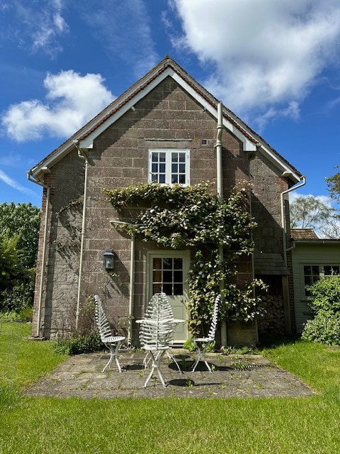 The exterior of the cottage features a stone façade adorned with greenery, offering a welcoming entrance. A patio area is presented with four white, slatted chairs arranged around a small table, all set against a backdrop of well-maintained lawn and bright blue skies.