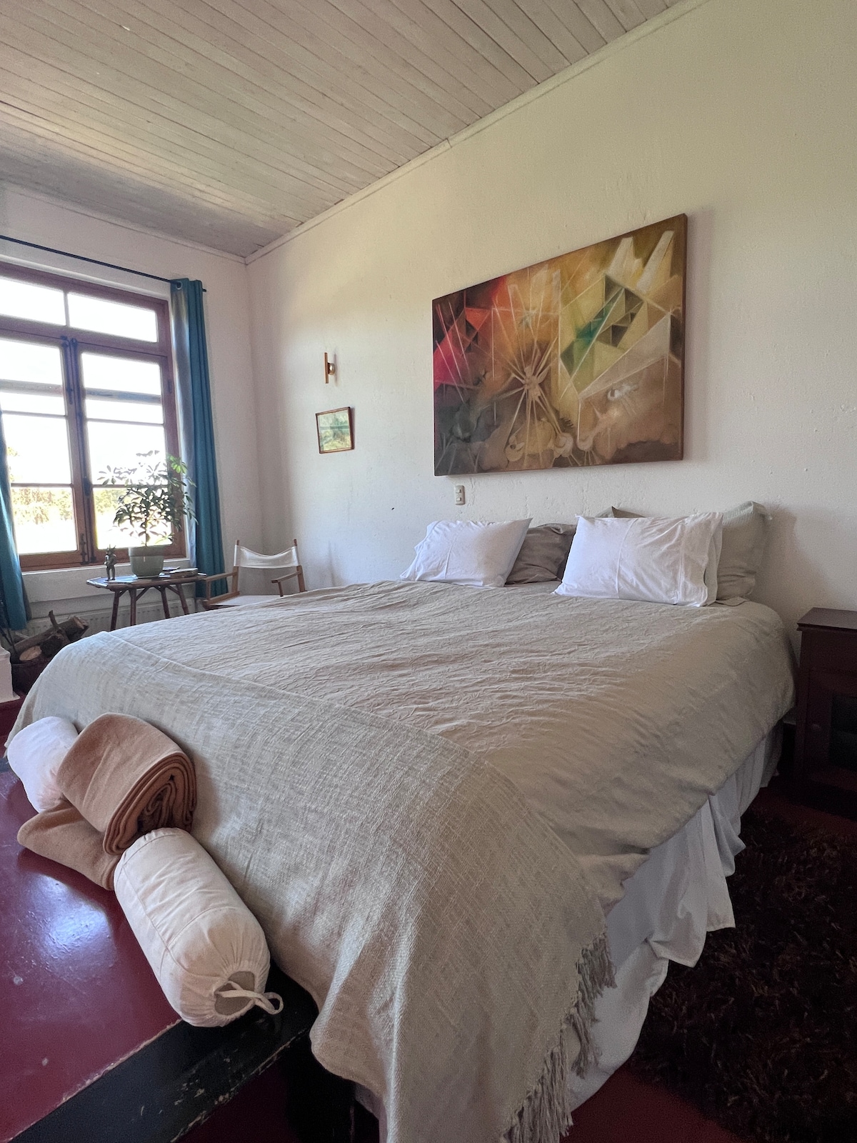 A spacious bedroom features a large bed dressed in neutral linens with multiple pillows. Colorful wall artadds visual interest above the bed. A window with sheer curtains allows natural light to fill the room, while a small chair and table are positioned nearby.