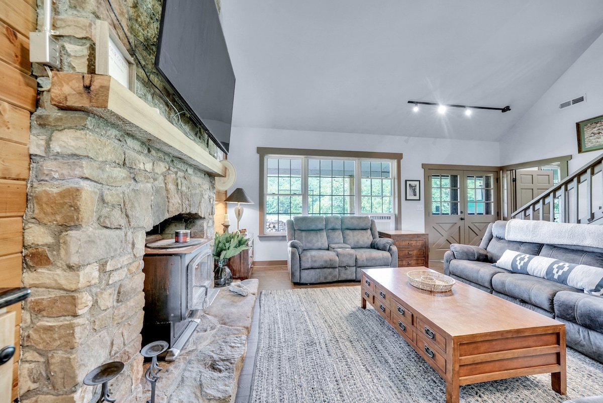 A spacious living area features a rustic stone fireplace, providing warmth and an inviting focal point. Comfortable seating options include two plush sofas positioned around a wooden coffee table. Large windows allow natural light to illuminate the room, creating a bright and open atmosphere.