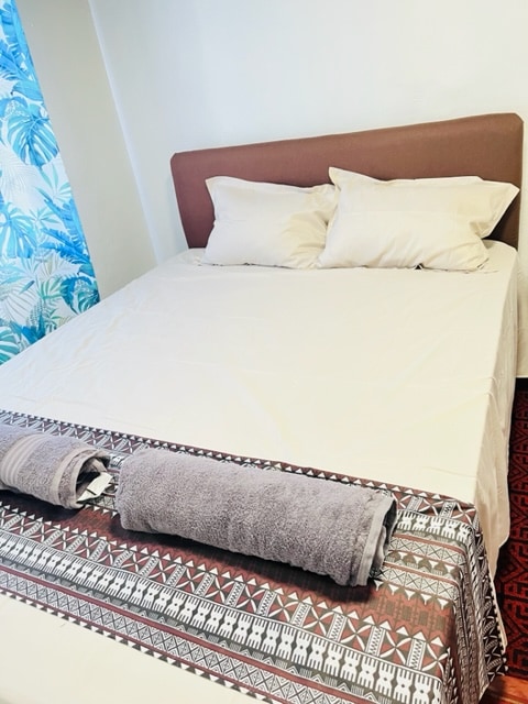 A spacious bed is presented with neatly arranged pillows and a fitted sheet. A decorative blanket rests at the foot of the bed. Natural light filters in through a window adorned with a tropical-themed curtain, adding a touch of brightness to the room.
