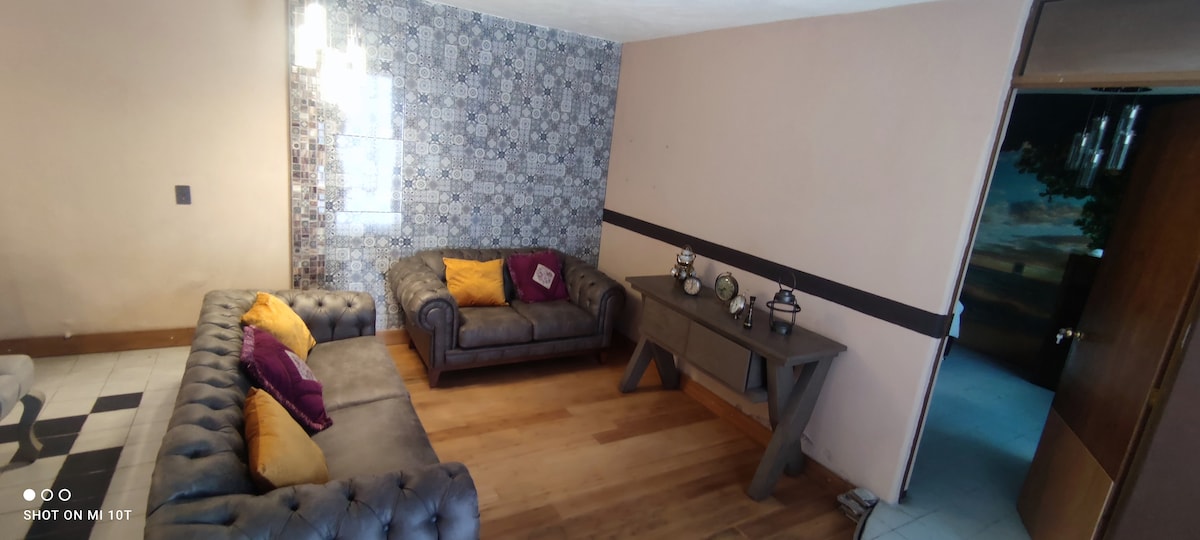 A comfortable living area features a gray chesterfield sofa with colorful cushions and a wooden table. Decorative wall tiles add texture, and natural wood floors create warmth. Soft light filters through an adjacent doorway leading to another space.