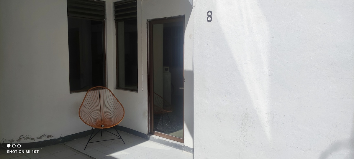 An outdoor area is presented with a modern chair positioned beside a door. Light-colored walls and large windows create an open feel. The floor is laid with simple tiles, and a glimpse of an indoor space is visible through the door.