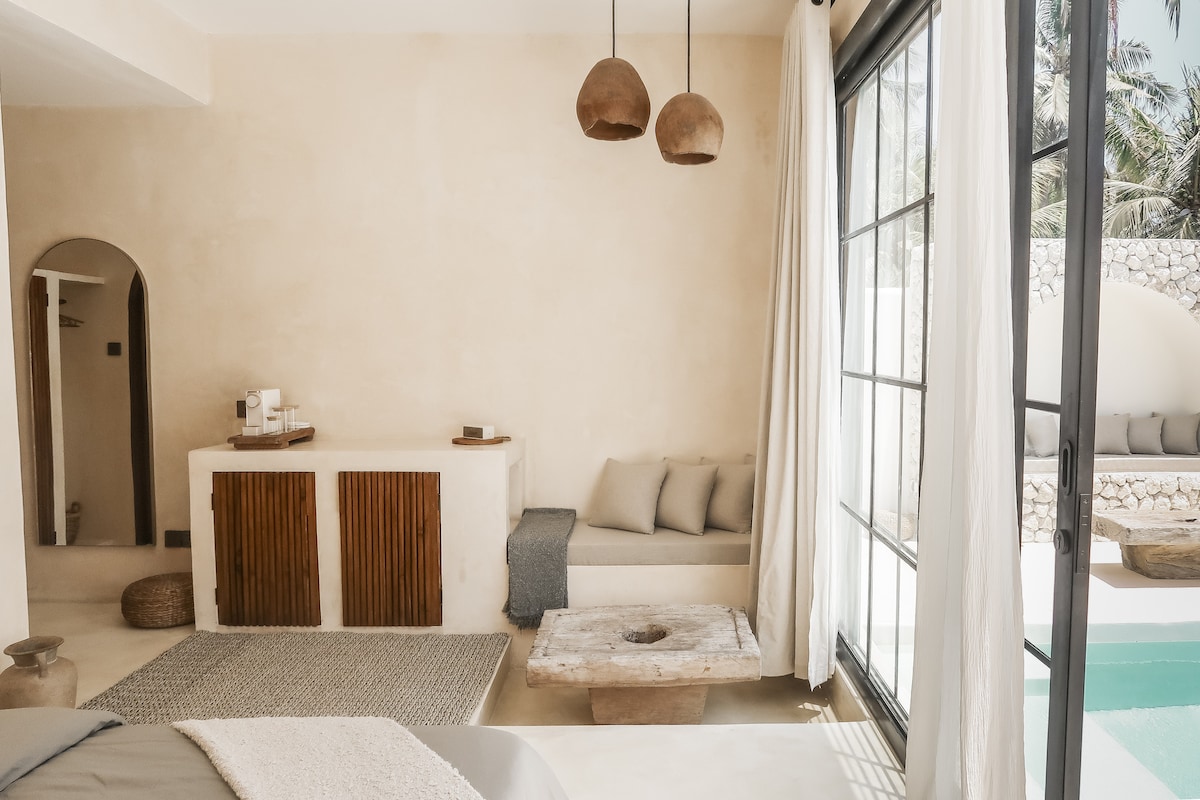 The interior features a neutral color palette with textured walls. A cozy sitting area with soft cushions is positioned next to large glass doors, providing a view of the pool. A minimalist wooden bar area is complemented by two pendant lights hanging above.