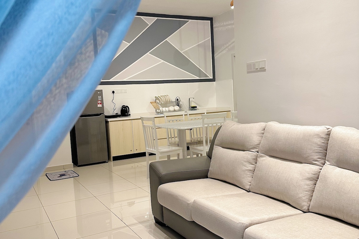 The living area showcases a modern, L-shaped sofa in neutral tones, inviting relaxation. A sleek dining table with chairs is visible in the background, and a kitchenette area features appliances including a kettle and refrigerator. Ambient lighting enhances the clean, tiled floor.