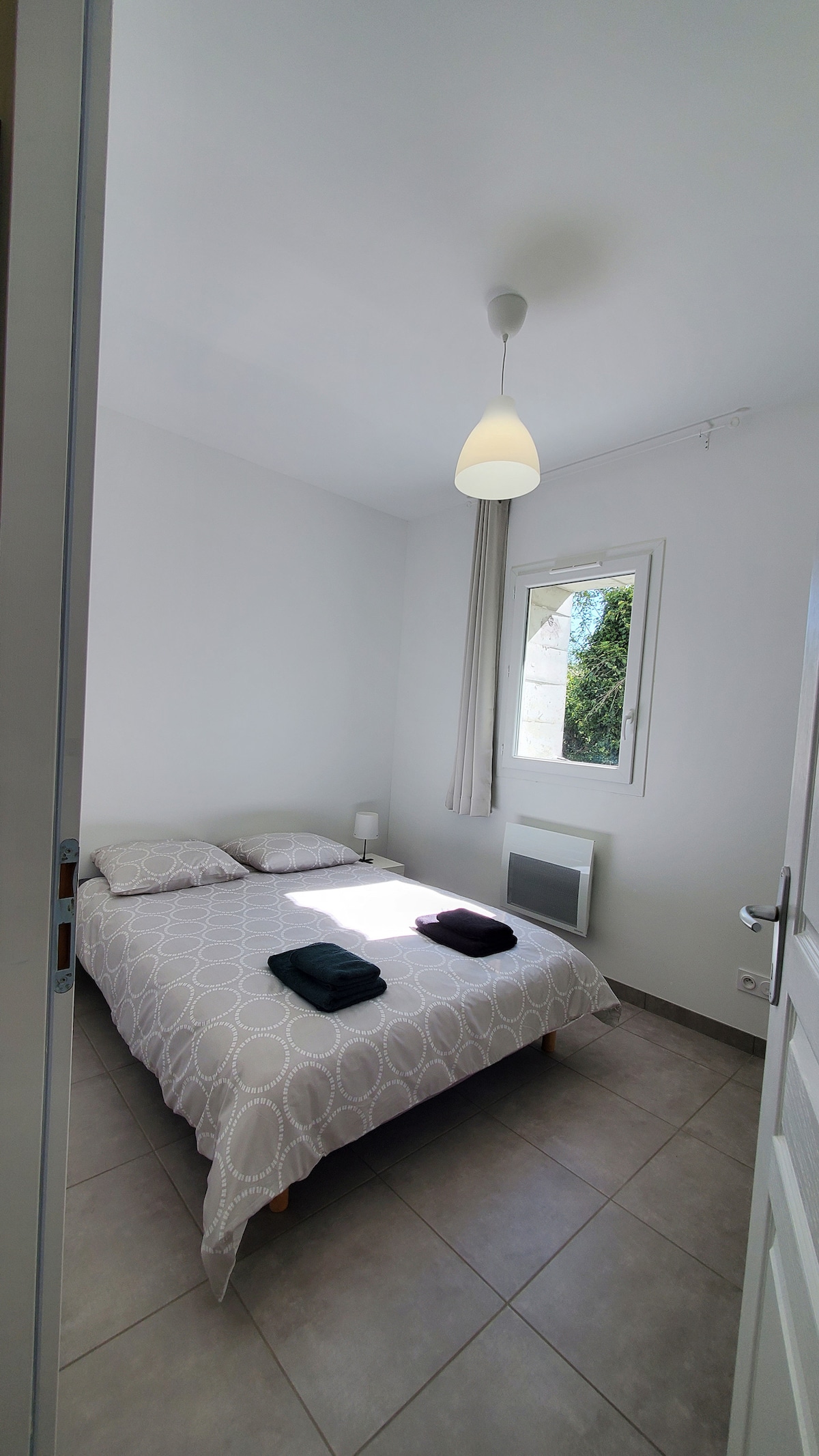 A bright sleeping area is furnished with a spacious bed and light bedding. Two bedside lamps are positioned next to the bed. A window allows natural light to enter the room, while a heater and a window are visible on the adjacent wall.