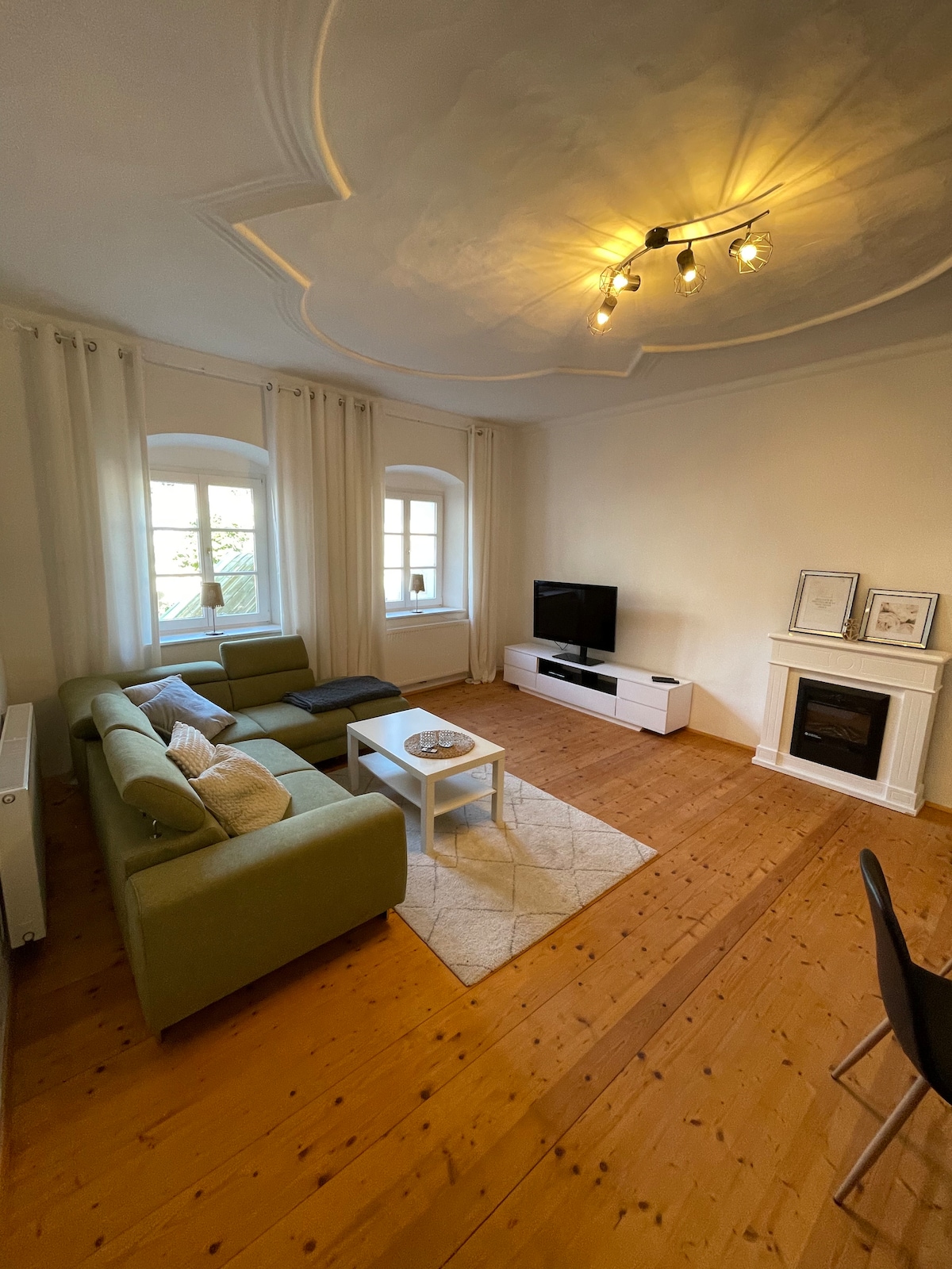 A living area presents a light-colored sofa facing a television stand. Natural light enters through two windows dressed with sheer curtains. A white coffee table is positioned centrally on a woven rug atop wooden flooring, accented by a decorative fireplace and overhead lighting.
