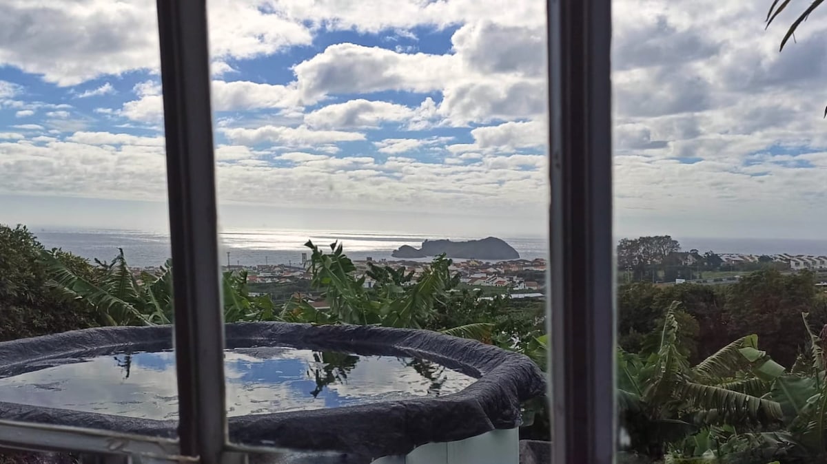 A serene view of the Atlantic Ocean is captured through a window frame. The surface of a calm pool reflects the sky, with a distant island visible on the horizon. Lush greenery surrounds the scene, enhancing the connection to nature.