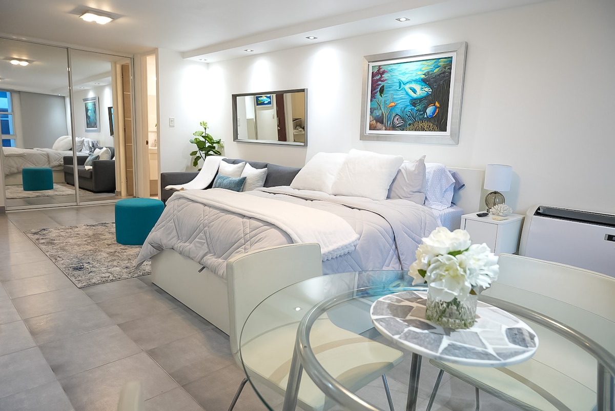A modern studio apartment is displayed featuring a queen bed with crisp linens. A cozy sofa sits nearby, and a glass dining table is set for two, surrounded by neutral tones. Wall mirrors enhance the light and space, while artwork adds a touch of color.