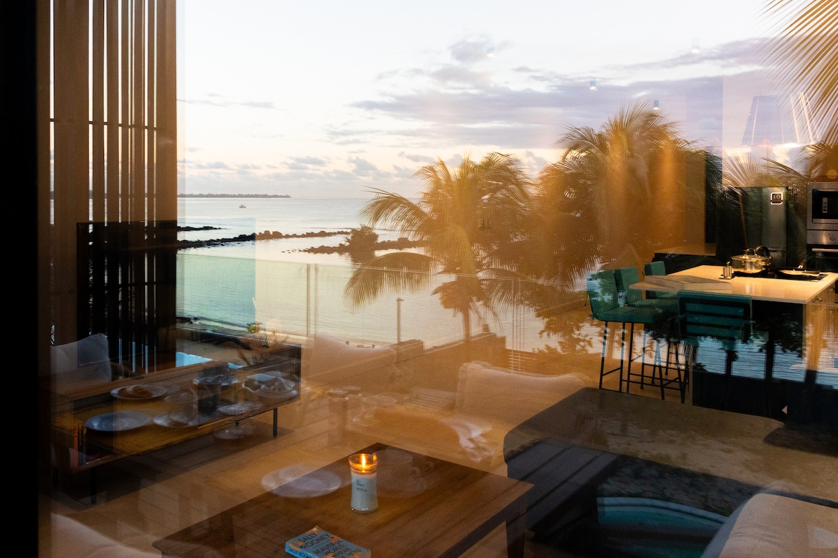 A serene vista is reflected in the glass panels, showcasing a tranquil waterfront view with palm trees swaying gently. A cozy seating area and a candlelit table create an inviting ambiance. The soft glow of sunset complements the peaceful environment.