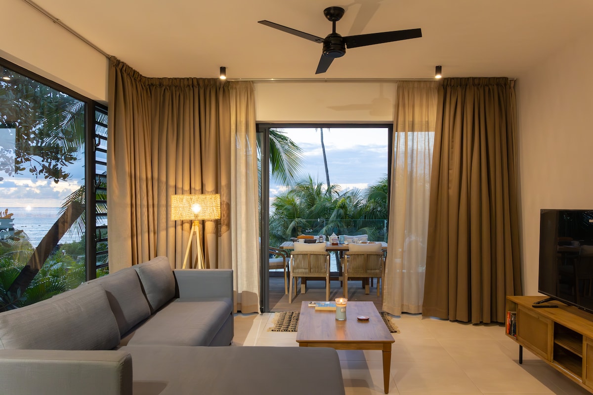 A spacious living area features large windows offering views of palm trees and the ocean. A comfortable sofa and a wooden coffee table are present, while a standing lamp provides soft lighting. Sheer and heavy curtains frame the windows, enhancing the serene ambiance.