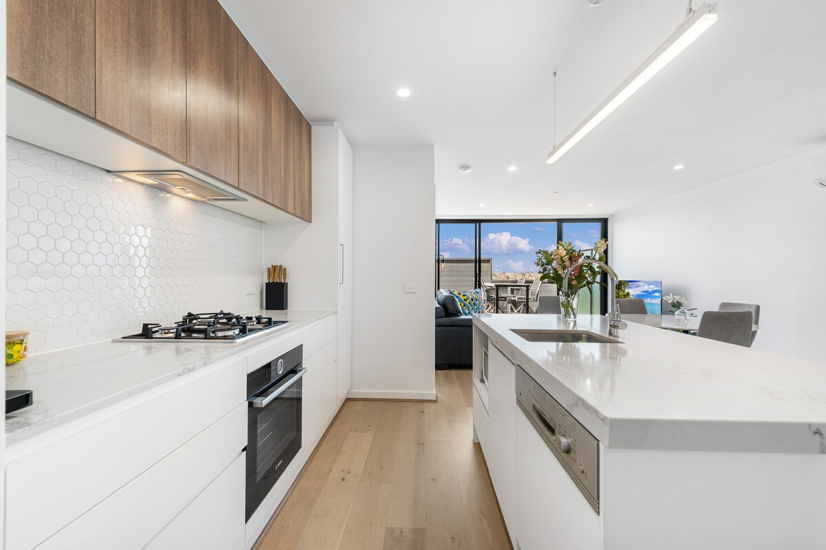A modern kitchen features a combination of light cabinetry and wooden accents. Stainless steel appliances, including a gas stove and oven, are integrated into the space. A sleek island countertop offers additional workspace, while large windows provide natural light and views of the living area.