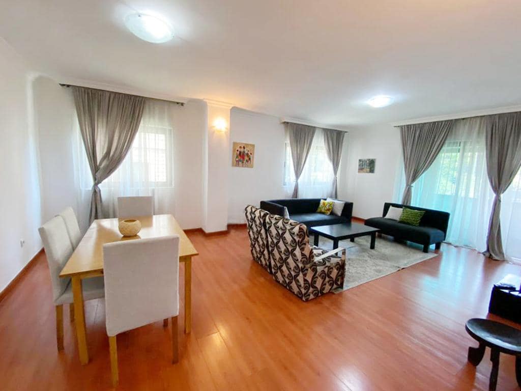 A bright living area featuring a combination of seating arrangements, including a patterned sofa and a dark couch. A light wood dining table with four chairs is present. Large windows are adorned with sheer curtains, allowing natural light to fill the space.
