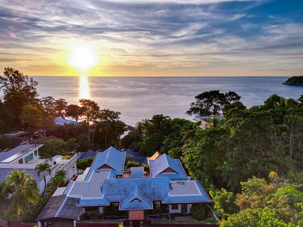 Aerial view of a villa set among lush greenery, with a stunning ocean backdrop. The sun sets over the horizon, casting a warm glow across the sky. The villa features a traditional design, with a multi-pitched roof and expansive outdoor space.