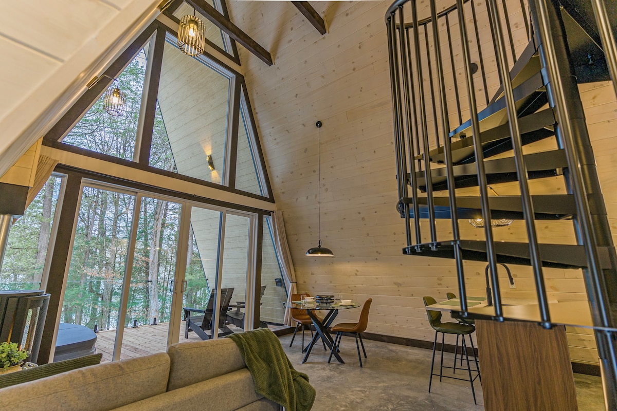 A modern interior features an open living space with high ceilings and large triangular windows. A spiral staircase leads to the upper level. A cozy couch is positioned near a wooden dining table with stylish chairs, while outdoor views are visible through the glass doors.