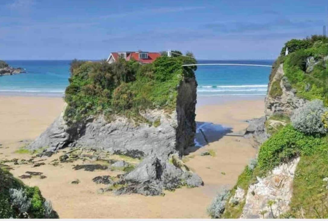 A rocky outcrop rises from a sandy beach, surrounded by greenery. The background features a calm sea with gentle waves, and a clear blue sky above, creating a relaxing coastal scene.