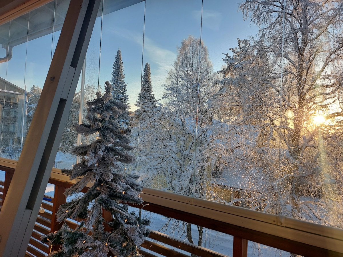 A furnished balcony offers a view of a winter landscape, featuring snow-covered trees and a soft sunrise illuminating the scene. A small decorative tree is placed on the balcony railing, enhancing the peaceful ambiance of the setting.